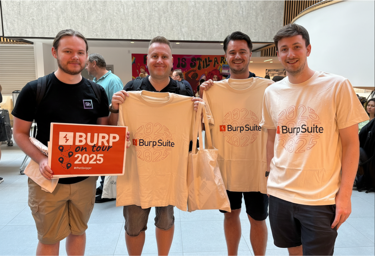 Four men standing indoors at an event, smiling and holding beige T-shirts printed with "Burp Suite 2025." One man also holds a sign reading "BURP on tour 2025 by PortSwigger." The group appears to be in a busy venue with other attendees in the background.