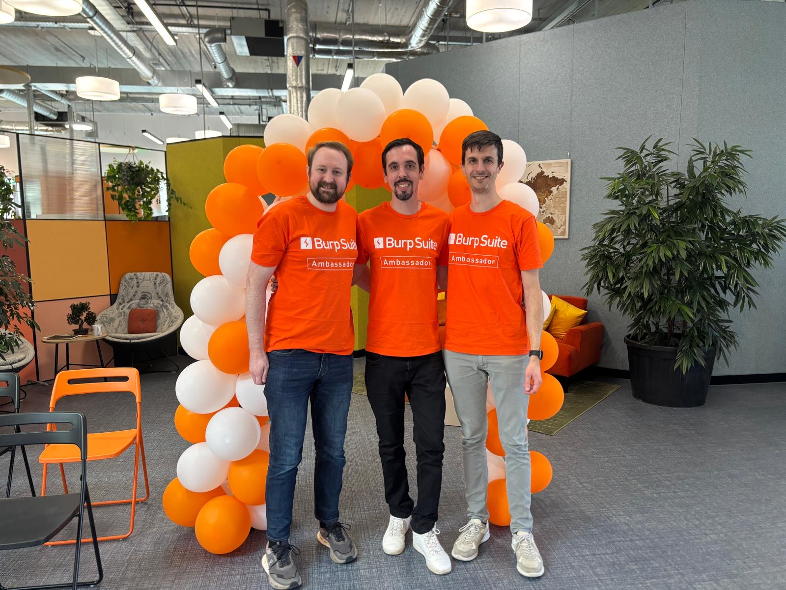 Tib3rius, El Mago, and Federico Dotta wearing bright orange "Burp Suite Ambassador" T-shirts stand together smiling in an office space, posing in front of an arch made of orange and white balloons. The background includes modern office furniture, plants, and exposed ceiling ducts.